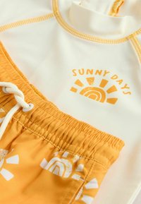 Fehér rash guard narancssárga varrással, "SUNNY DAYS" felirattal. Sárga fürdőnadrág fehér nap- és szivárványmintákkal.