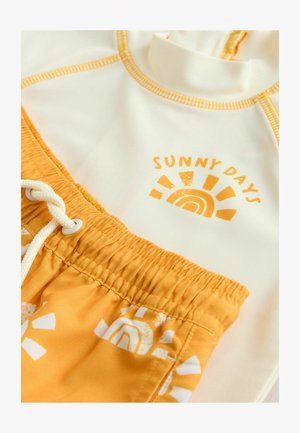 Biely rash guard s oranžovým prešívaním a grafikou "SLNEČNÉ DNI". Žlté plavkové šortky s bielymi vzormi slnka a dúhy.
