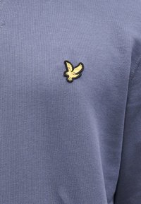 Sinine puuvillane särk, millel on rinnal väike musta ja kuldse kotka tikand ning pehme, tekstuuriga viimistlus.