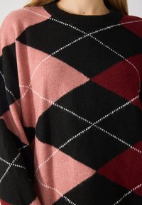 Pull en tricot avec un motif en losanges en rose, noir et bordeaux. Présente un col rond et des coutures blanches le long des bords des losanges.