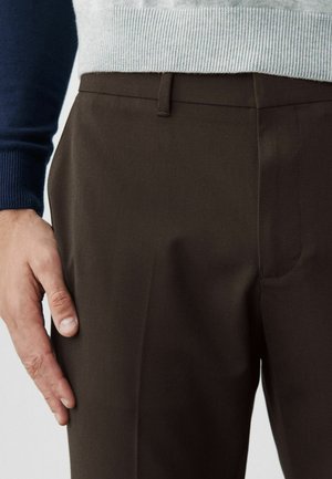 Hnědé kalhoty s hladkou texturou a přizpůsobeným střihem, s jednou přední kapsou, zobrazené vedle šedého pleteného svetru.