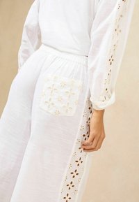 Tenue en tissu blanc avec des manches longues et une taille smockée, ornée de détails floraux brodés sur le côté et la poche arrière.