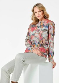 Langärmlige Bluse im mehrfarbigen Blumenmuster mit lebhaften Nuancen von Pink, Blau und Beige. Kombiniert mit hellgrauen Hosen.