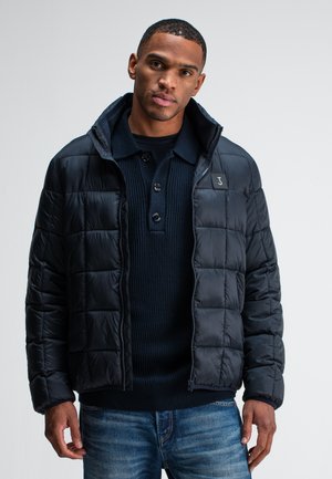 Schwarze gefütterte Jacke mit einem gesteppten Design und Reißverschluss, kombiniert mit einem marineblauen ribbeligen Strickpullover. Das Model posiert vor einem schlichten Hintergrund.
