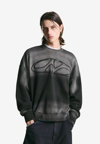 Jongeman met nat-uitziend haar van gemiddelde lengte, draagt een vervaagde zwarte sweatshirt met een abstract logo, handen in de zakken van donkere broek.