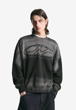 Jongeman met nat-uitziend haar van gemiddelde lengte, draagt een vervaagde zwarte sweatshirt met een abstract logo, handen in de zakken van donkere broek.