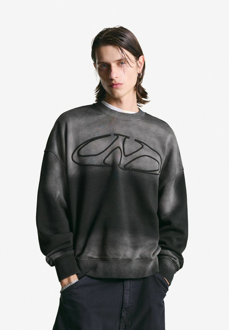 Jongeman met nat-uitziend haar van gemiddelde lengte, draagt een vervaagde zwarte sweatshirt met een abstract logo, handen in de zakken van donkere broek.