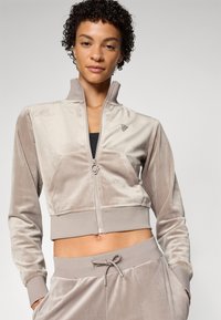 Velvet zip-up jacket in light beige, cropped design, featuring a stand-up collar, front pockets, and smooth texture. Accented with a small logo.