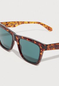 Γυαλιά ηλίου με σχέδιο χελώνας, πράσινους φακούς και τετράγωνα πλαίσια. Οι βραχίονες από υφή πλαστικού διαθέτουν αντίθετο μοτίβο και λογότυπο.