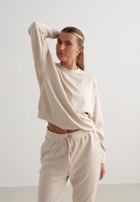 Felpa corta beige con scollo rotondo, maniche lunghe e dettaglio del logo sul petto, abbinata a pantaloni jogger beige coordinati con vita elastica.