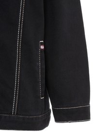 Schwarze Denimjacke mit kontrastierendem beigem Stich, einer Seitentasche mit Logo-Tag und abgerundetem Saum. Die Oberfläche ist glatt mit sichtbaren Nähten.