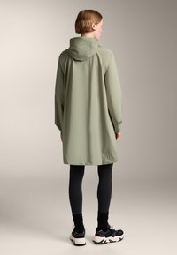 Világoszöld, vízálló, kapucnis esőkabát, térd hosszúságú, laza szabással és varrási részletekkel, fekete leggings-szel és sportcipővel párosítva.