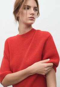 Pull rouge en tricot avec une texture côtelée, manches courtes et col rond. Le modèle est debout devant un fond blanc uni, les bras croisés.