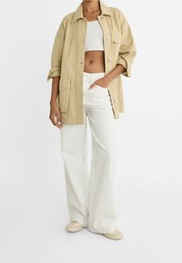 Béžová bavlněná košile s dlouhým rukávem a dvěma předními kapsami, nošená přes bílý zkrácený top a bílé kalhoty širokého střihu. Boty jsou světle béžové.