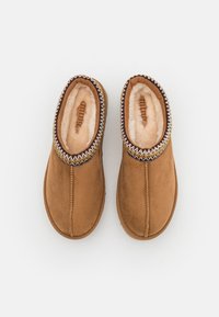 Chaussons en daim marron avec une doublure douce de couleur claire et un détail de couture texturé autour du col. Bout arrondi et semelle plate.