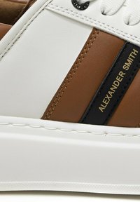 Sneaker in pelle bianca e marrone con accenti neri, caratterizzata da una texture liscia, design cucito e dettaglio di branding dorato.