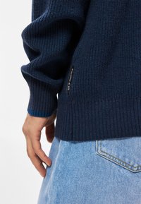 Mørkeblå strikket sweater med ribstrikkede manchetter og kant, der har en glat tekstur og et lille mærke på siden med teksten "EST. IN STOCKHOLM."