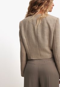 Bēša krītoša bleizeris ar teksturētu apdari, kuram ir vienkāršs kakla izgriezums un strukturētas plecu daļas, apvienots ar piegrieztiem brūniem biksēm.