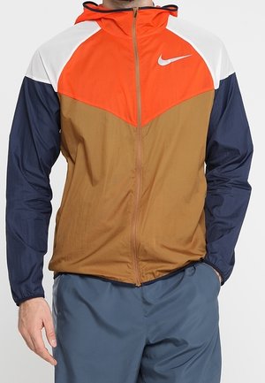 Mann trägt eine colorblock Nike-Jacke mit orange, braun, marineblau und weißen Bereichen und blaue Hose, steht mit den Händen neben dem Körper.