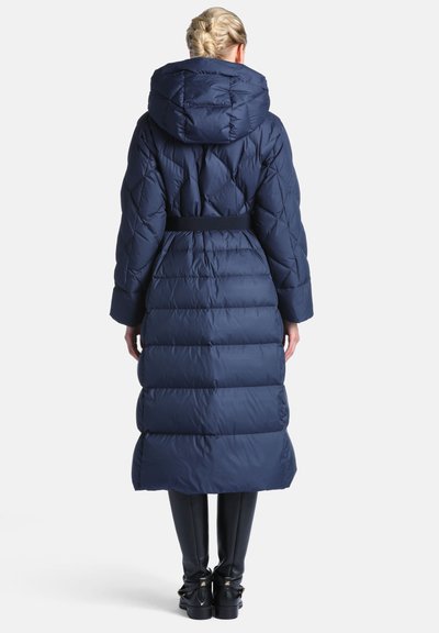 Femme aux cheveux tressés porte un long manteau matelassé bleu marine avec capuche, ceinture noire, pantalon noir et bottes noires, debout de dos.