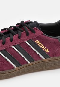 Bordeauxfarbene Wildledersneaker mit drei schwarzen Streifen, goldenem "SPEZIAL"-Logo und strukturierten Gummisohle. Verfügt über schwarze Akzente und Schnürdesign.