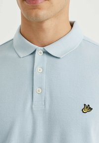Polo shirt bleu clair avec un tissage texturé, quatre boutons blancs et un petit logo brodé jaune sur le côté gauche de la poitrine.