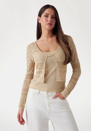 Beige knit cardigan with textured design and long sleeves, layered over a matching top, paired with white pants. Notable patterns on sleeves.