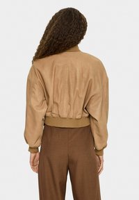 Frau mit lockigem Haar trägt eine kurz geschnittene, beige Jacke mit gerippten Manschetten und Saum, kombiniert mit hoch taillierten braunen Hosen, von hinten gezeigt.