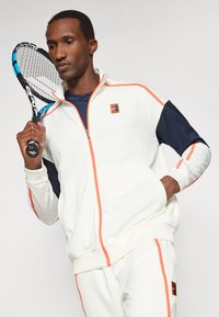 Λευκό αθλητικό μπουφάν με ναυτικές μανίκες και κοραλλί λεπτομέρειες, διαθέτει φερμουάρ και λογότυπο, συνδυασμένο με παντελόνι τζόγκινγκ αντίστοιχου χρώματος.