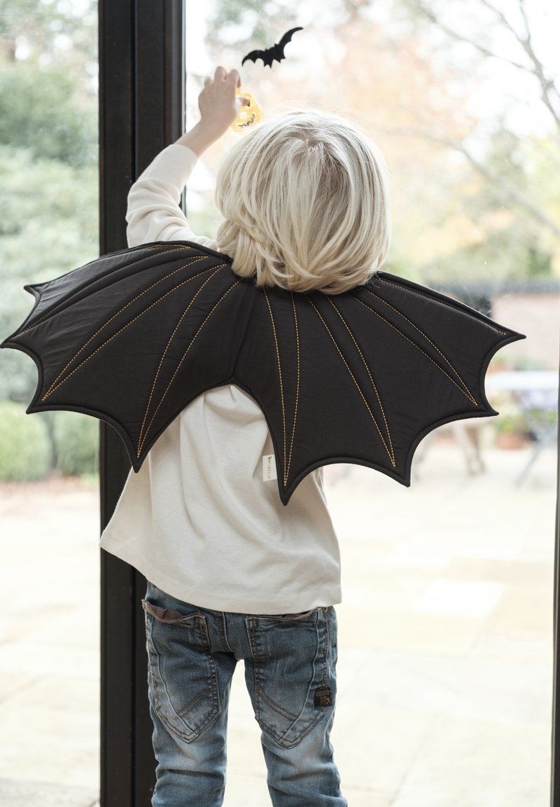 Aripi negre în formă de liliac cu cusături galbene, purtate pe umeri, atașate unei cămăși albe cu mâneci lungi și blugi albastre ai unui copil.