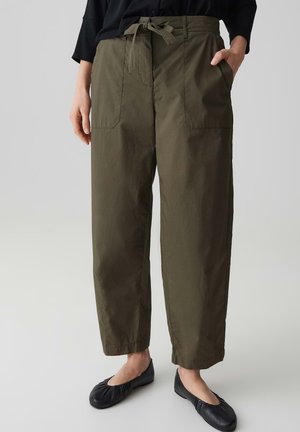 Femme portant un pantalon taille haute vert olive avec ceinture nouée, grandes poches avant, associé à des ballerines noires et un haut noir.