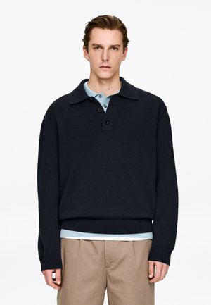 Dark navy knitted pullover with a wide collar and three-button placket, layered over a light blue shirt, paired with beige trousers.