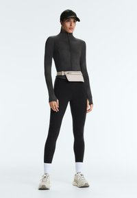 Femme portant une veste noire à fermeture éclair, des leggings noirs, des baskets blanches, des chaussettes blanches, une casquette noire et une pochette beige à la taille, debout devant un fond uni.