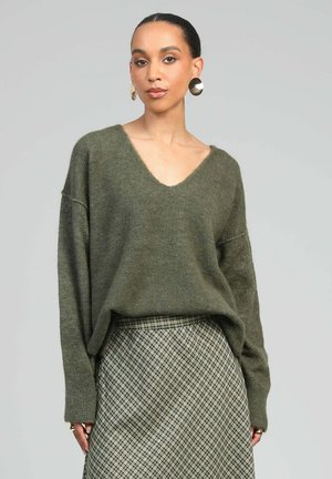Femme portant un pull col en V oversize couleur vert olive et une jupe taille haute à carreaux beige et noire sur un fond gris uni.