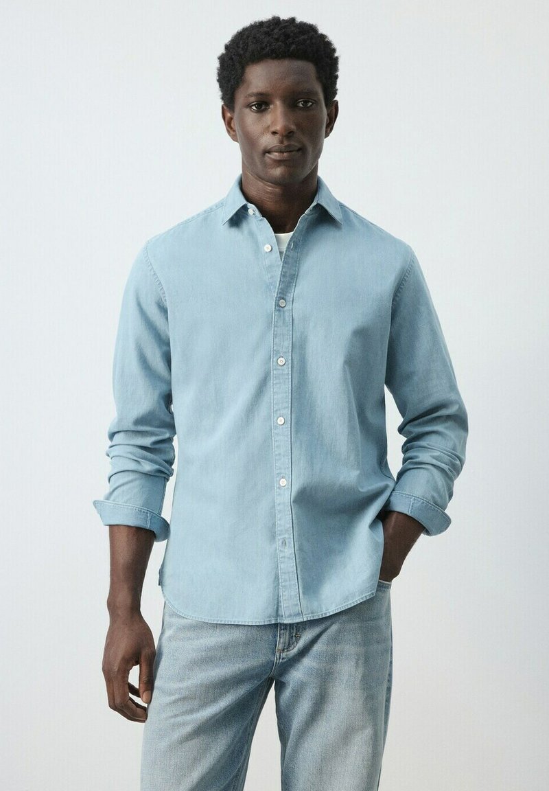 Homme portant une chemise en denim bleu clair avec les manches retroussées et un jean assorti bleu clair, debout devant un fond blanc uni.