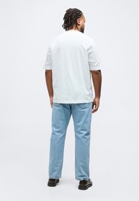 Άνδρας με πλεξούδες που φοράει απλό λευκό μπλουζάκι, ανοιχτόχρωμα μπλε τζιν και μαύρα παπούτσια, στέκεται με την πλάτη σε λευκό φόντο.