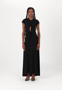 Abito maxi nero con dettaglio a nodo sul décolleté, maniche corte e accentuazione cut-out. Tessuto liscio con una leggera lucentezza, indossato con tacchi neri.