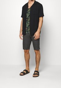 Camisa negra de manga corta y botones sobre una camiseta de tirantes verde con flores, combinada con pantalones cortos grises y sandalias negras con hebillas.