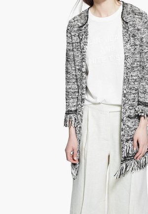 Cardigan a maglia in bianco e nero con dettagli in frange, abbinato a una t-shirt bianca con stampa testuale e pantaloni larghi color crema.
