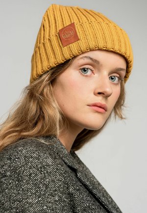 Żółta dziana czapka z żebrową teksturą i kwadratową skórzaną łatką. Modelka ma na sobie szary żakiet w jodełkę, a jej włosy mają miękkie fale.