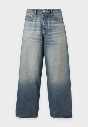 Mann mit dunkler Hautfarbe, der ein lockeres, ausgewaschenes schwarzes T-Shirt mit Schriftzug und weite, ausgewaschene blaue Jeans trägt, steht vor einem schlichten grauen Hintergrund.