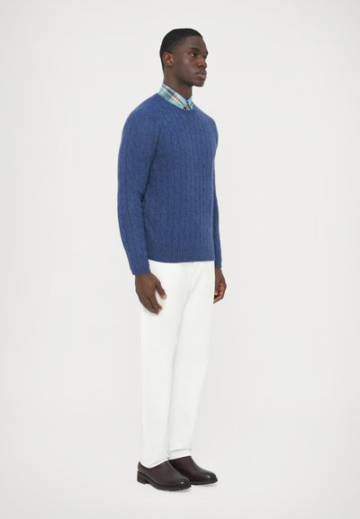 Mėlynas megztinis su pynimo raštu, pagamintas iš vilnos, dėvimas virš languoto marškinių su apykakle. Derinamas su baltais kelnėmis ir rudais slip-on tipo batais.