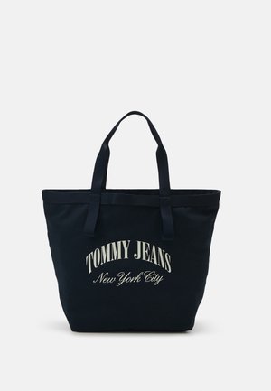 Czarna torba na ramię z podwójnymi uchwytami, z białym napisem "Tommy Jeans New York City" z przodu, na jednolitym jasnym tle.