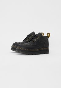Botas de cuero negro con detalles de costura, punteras redondas, acentos amarillos y suela de goma gruesa. Cuenta con una lengüeta en el talón.