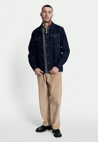 Mann i mørkeblå jakke, grønn skjorte, beige kordfløyelsbukser, svarte sko og mønstret halsduk foran hvit bakgrunn.