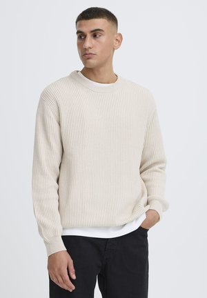 Mand med kort hår iført en cremefarvet ribstrikket sweater over en hvid skjorte og sorte bukser, kigger let mod venstre.