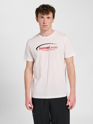 Junger Mann mit lockigem Haar, der ein weißes Hummel Sport T-Shirt und schwarze Hose trägt, steht vor einem schlichten, hellen Hintergrund.