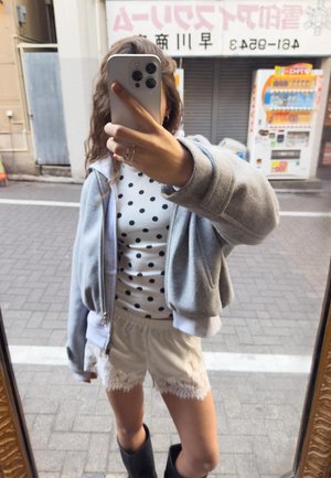 Persona con giacca grigia, camicia bianca a pois, pantaloncini in pizzo e stivali neri che scatta un selfie allo specchio su un marciapiede della città.