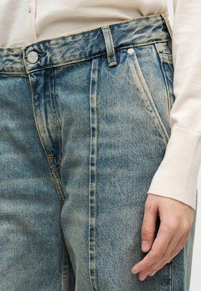 Κοντινό πλάνο ενός ατόμου που φοράει ξεθωριασμένο μπλε τζιν και κρεμ πουκάμισο με μακριά μανίκια, με το δεξί χέρι χαλαρό δίπλα στο σώμα.