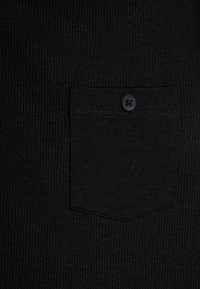 Schwarzer gerippter Stoff mit einer kleinen Brusttasche, die einen einzelnen schwarzen Knopf aufweist. Die strukturierte Oberfläche zeigt subtile Stoffdetails.